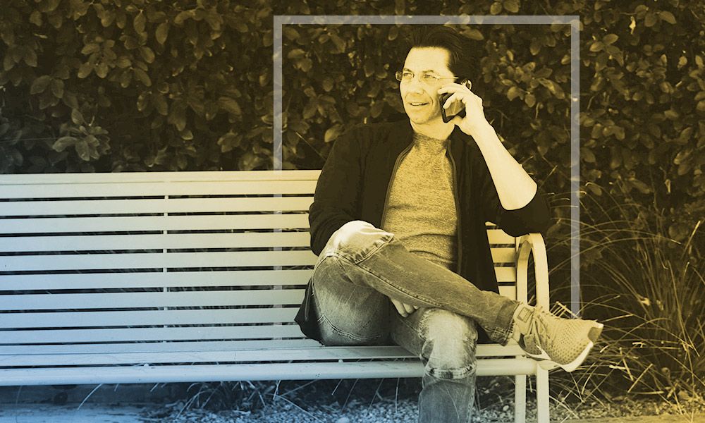 Dean Graziosi talking on his cell phone on a park bench, smiling at the conversation.